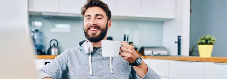 Man sitting in a kitchen, smiling and holding a coffee mug, wearing headphones around his neck.