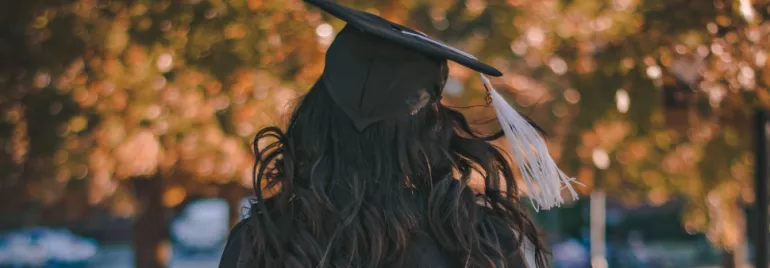Graduate in cap and gown standing with blurred autumn leaves in the background.