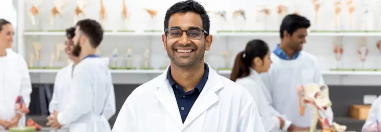 Smiling man in a lab coat stands in a scientific classroom with anatomical models in the background.