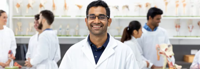 Smiling man in a lab coat stands in a scientific classroom with anatomical models in the background.