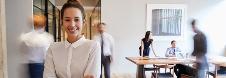 Smiling woman in white shirt stands with blurry office workers in background.