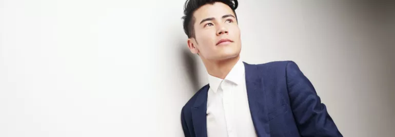 Man in blue suit looking up, leaning against a white wall.