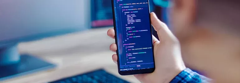 Person holding smartphone with code on screen, working at a computer.