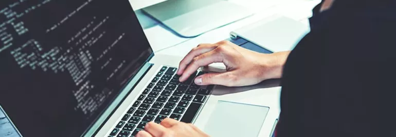 Person typing code on a laptop with a monitor showing code.