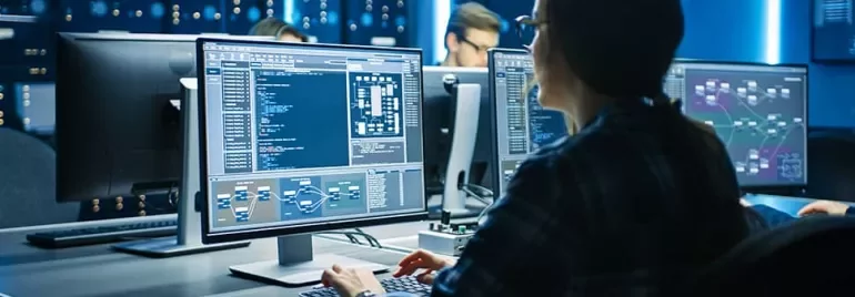 Person analyzing data on multiple computer monitors in a tech office.