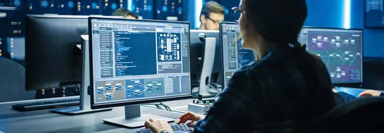 Person analyzing data on multiple computer monitors in a tech office.