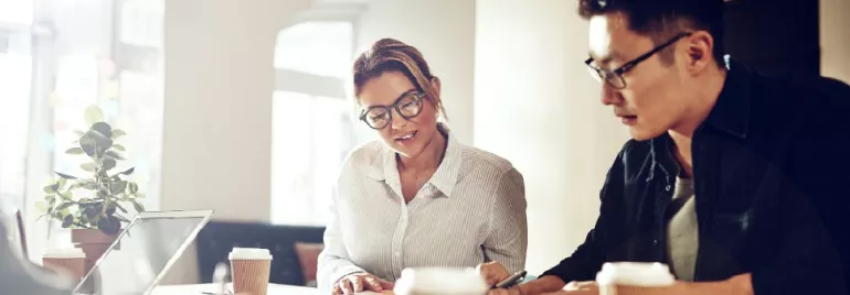 Two people in an office setting, collaborating with laptops and coffee cups on the table.