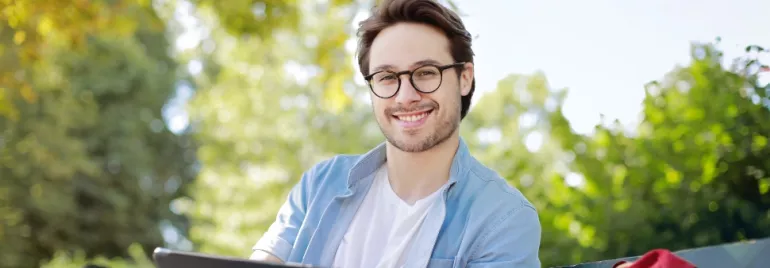 Person with glasses smiling outdoors on a sunny day.
