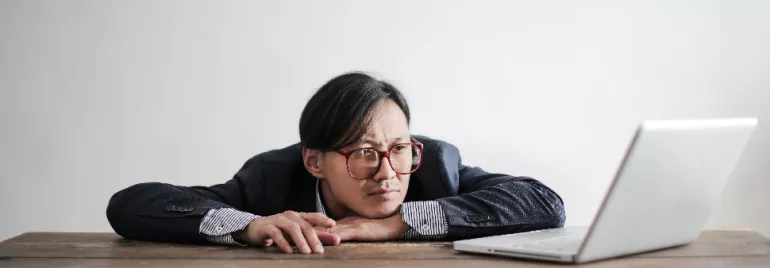 Man in glasses staring thoughtfully at a laptop on a wooden table.