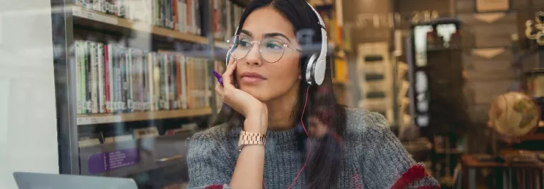 Woman with glasses and headphones, sitting in a library, looking thoughtful.