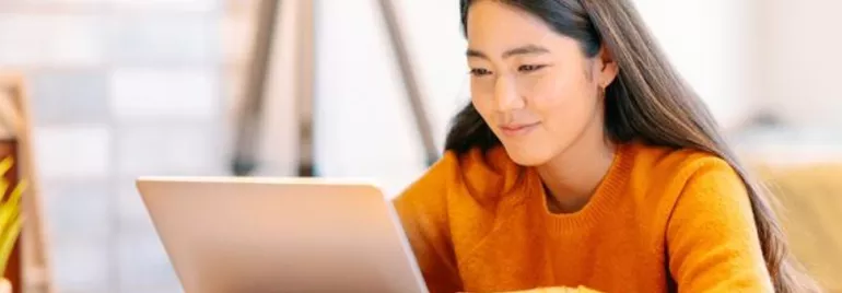 Woman in orange sweater using a laptop, seated indoors.