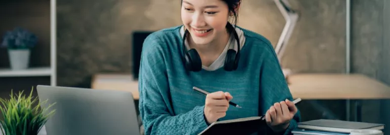 Woman in blue sweater studying with laptop and notebook.