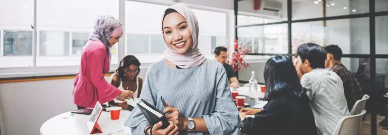 Smiling woman in office attire holding a notebook, with colleagues collaborating in the background.