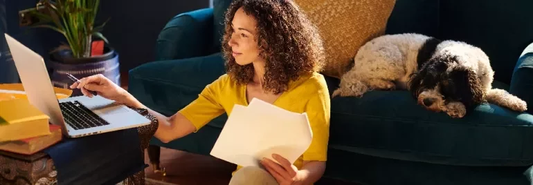 Woman in yellow shirt working on laptop with papers, dog resting on couch.