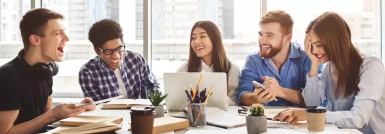 Group of five young adults laughing and talking while working together at a table, with a laptop and coffee cups.