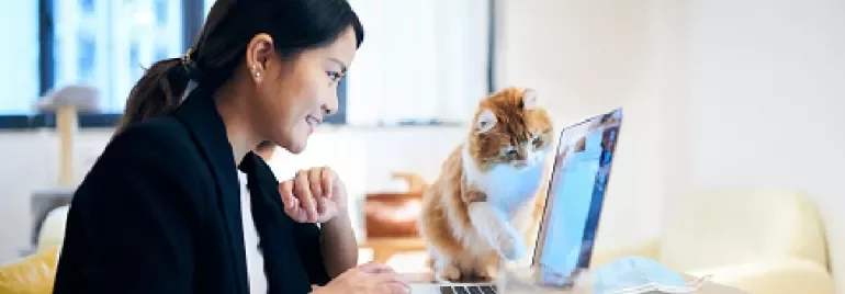 Woman smiling at laptop with curious cat beside her.