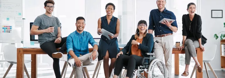 A diverse group of colleagues in an office, smiling and holding devices, with one person in a wheelchair.