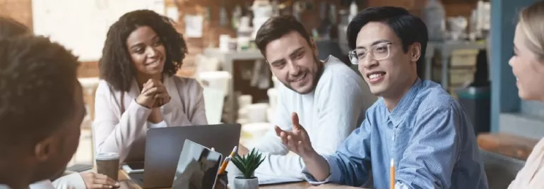 Group of people smiling and discussing at a meeting table with laptops and coffee cups.