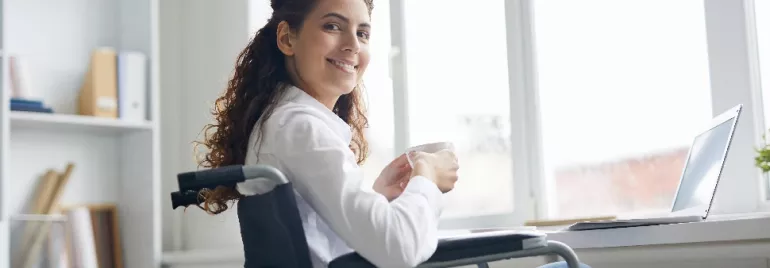 Woman in wheelchair smiling while holding a coffee cup at her desk.