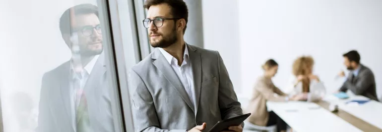 Man in a suit holding a tablet, standing by a window, with colleagues working in the background.