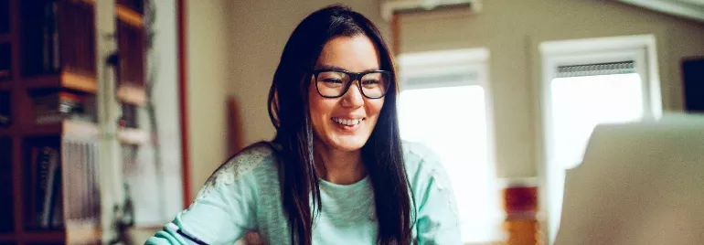 Woman in glasses smiling at a computer screen in a cozy room.
