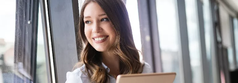 Smiling woman with long hair holding a tablet by a window.