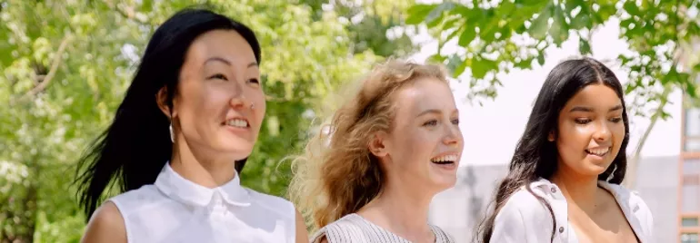 Three women walking outdoors, smiling under trees.