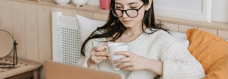 Woman wearing glasses, holding a mug, and using a laptop.