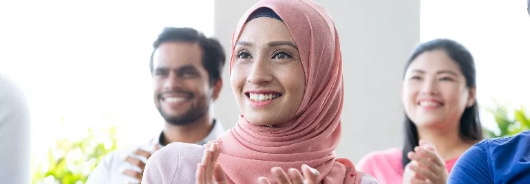 A smiling woman in a pink hijab applauds with others blurred in the background.