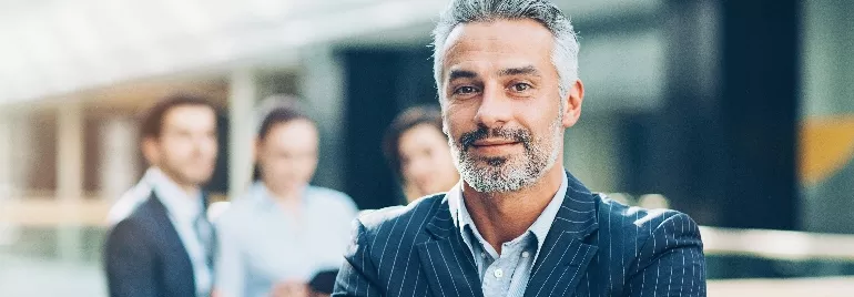 Confident businessman in a suit standing with a blurred office team in the background.