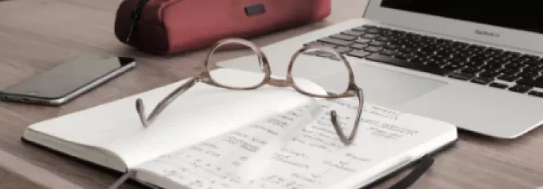 Notebook with eyeglasses on a desk next to a laptop and pencil case.