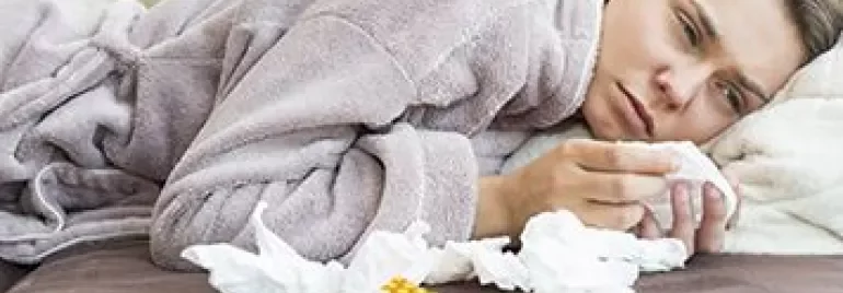 Person lying on a sofa in a robe, surrounded by tissues and medication.