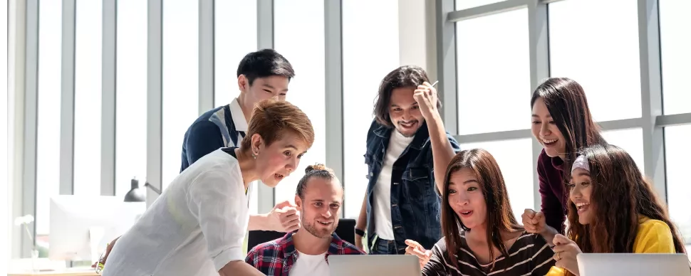 A group of people gathered around a laptop, looking surprised and delighted in a bright office setting.