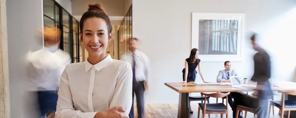 Smiling woman in white shirt stands with blurry office workers in background.