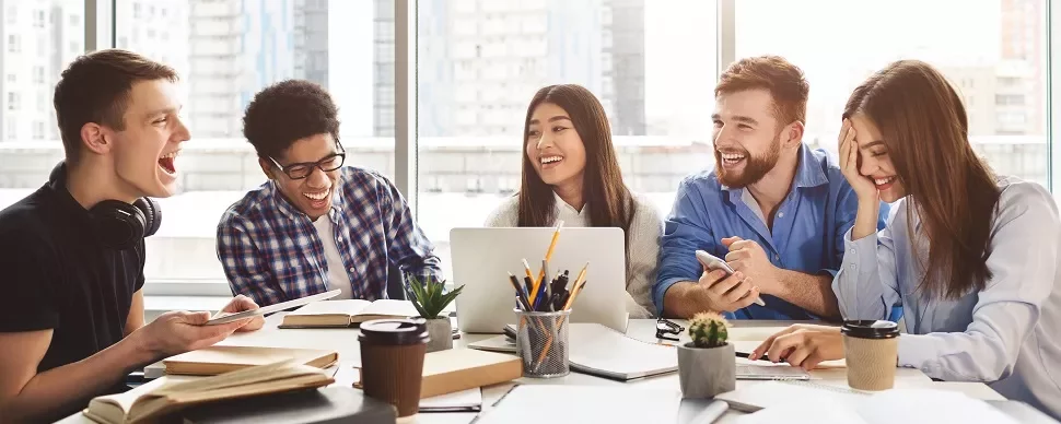 Group of five young adults laughing and talking while working together at a table, with a laptop and coffee cups.