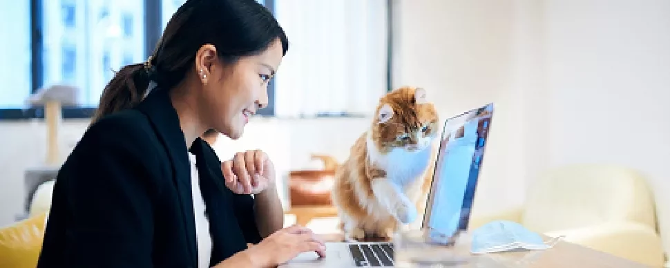 Woman smiling at laptop with curious cat beside her.