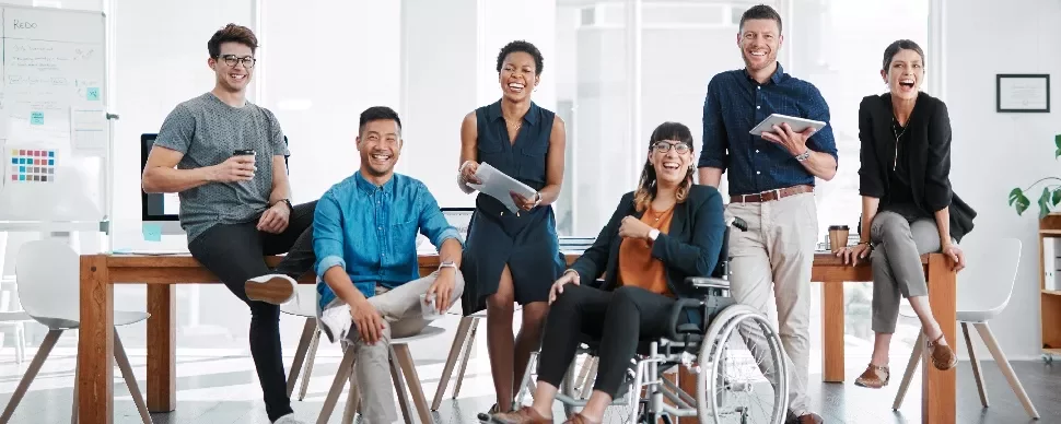 A diverse group of colleagues in an office, smiling and holding devices, with one person in a wheelchair.
