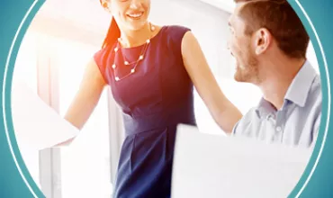 Woman in a blue dress smiles at a seated man with papers in an office setting.
