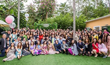 Large group of people posing outdoors on a lawn with trees and balloons.
