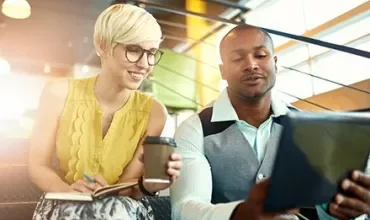 Two people discussing work over coffee, sitting on stairs with a tablet.