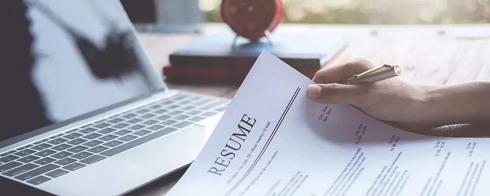 Person holding a resume in front of a laptop.