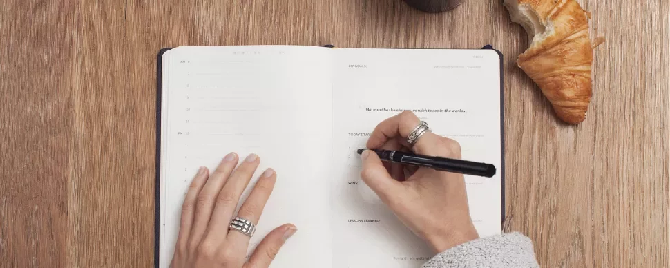 Person writing in a planner next to a croissant on a wooden table.