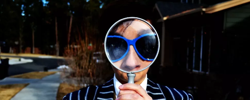 Person in sunglasses holding a magnifying glass to their face outdoors.