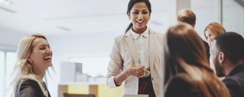 A woman in a suit smiling and talking to colleagues around a table in an office setting.