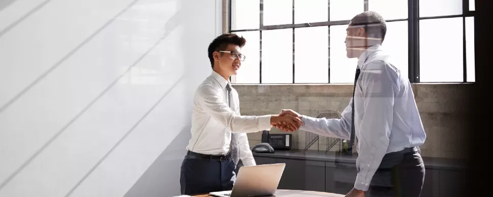 Two men in an office shaking hands, one with glasses and both in business attire.
