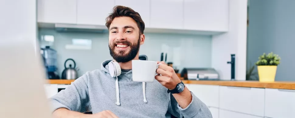 Man sitting in a kitchen, smiling and holding a coffee mug, wearing headphones around his neck.
