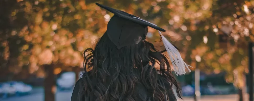 Graduate in cap and gown standing with blurred autumn leaves in the background.