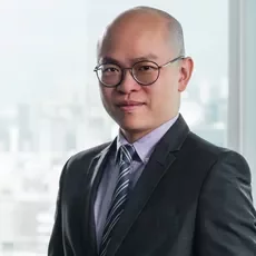 Smiling man in suit with glasses, standing indoors with a city skyline in the background.