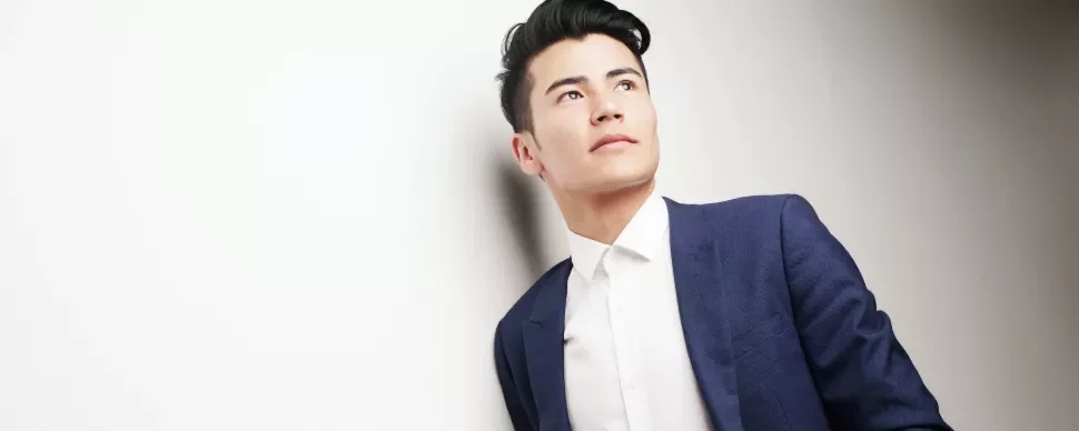 Man in blue suit looking up, leaning against a white wall.