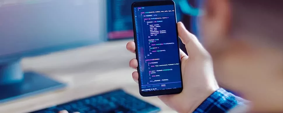 Person holding smartphone with code on screen, working at a computer.